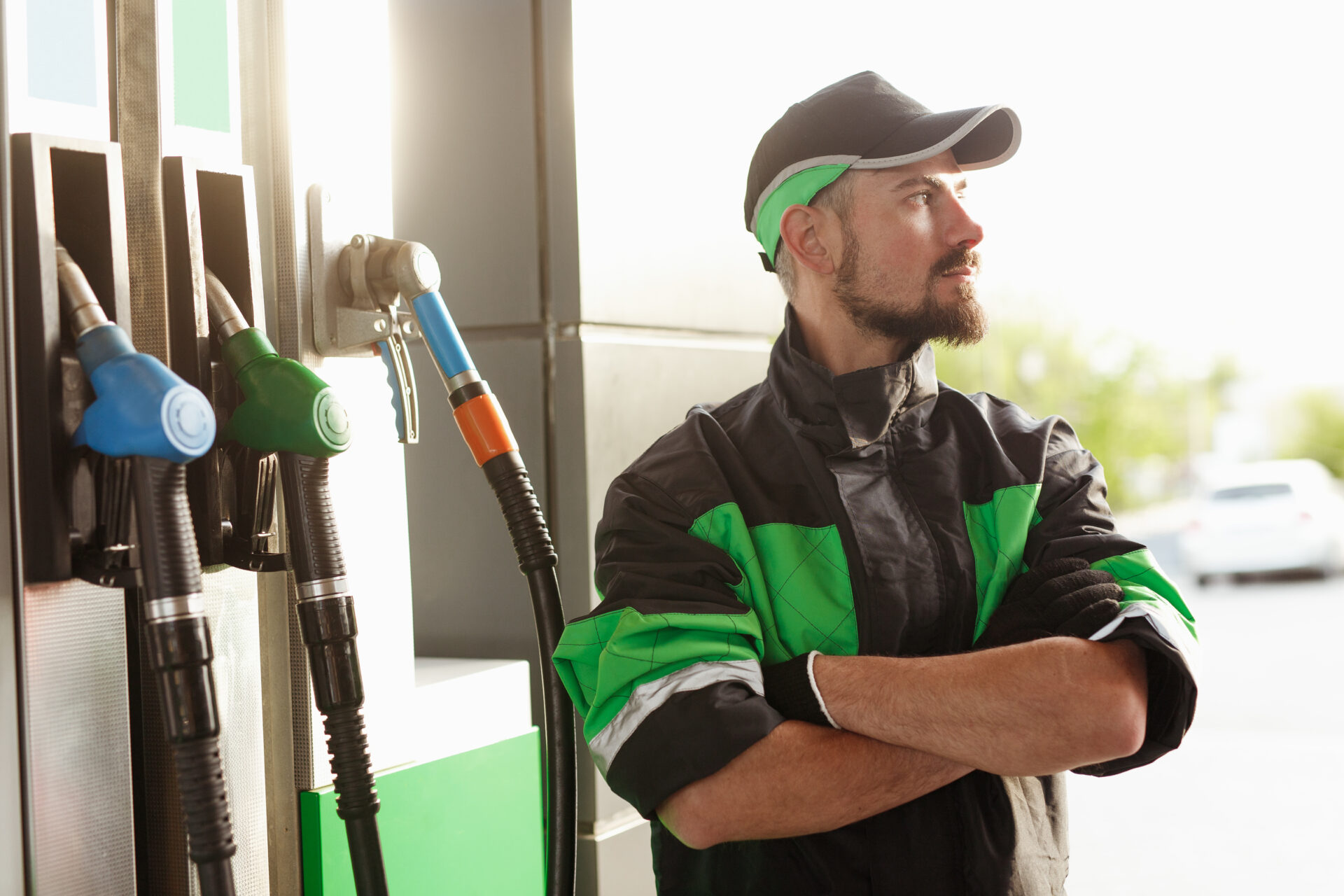 Self assured bearded man in uniform crossing arms and looking away while standing near petrol pump during work on gas station